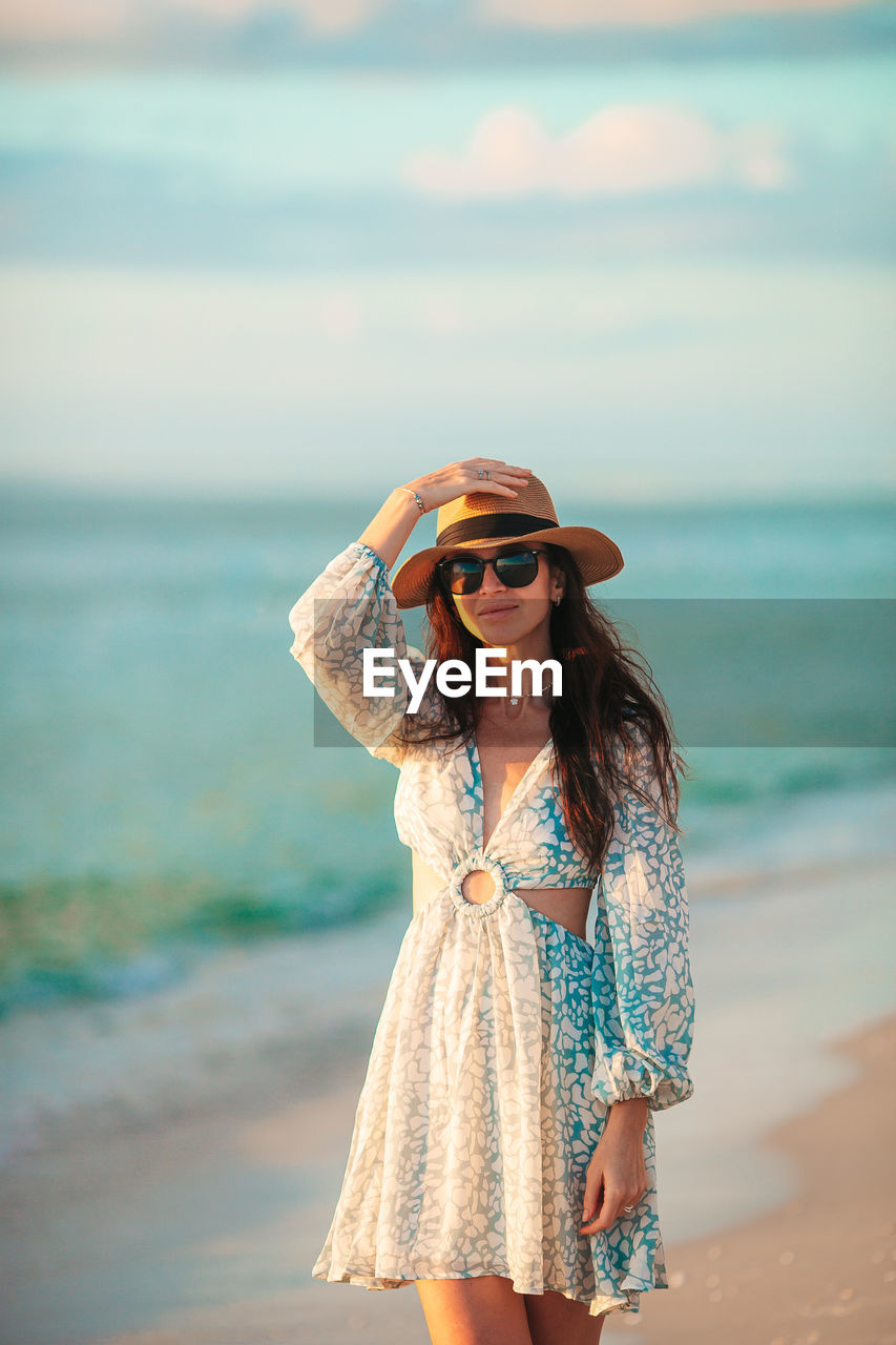 sea, water, fashion, beach, one person, land, clothing, women, adult, blue, young adult, sunglasses, dress, vacation, trip, nature, hat, holiday, sky, long hair, hairstyle, glasses, summer, standing, smiling, spring, leisure activity, happiness, portrait, photo shoot, travel, beauty in nature, brown hair, three quarter length, sand, travel destinations, casual clothing, horizon over water, emotion, front view, outdoors, female, horizon, lifestyles, day, person, relaxation, tourism, sun hat, focus on foreground, sunlight, looking, ocean, enjoyment, looking at camera, carefree, cheerful, copy space, cloud, teeth, smile, sunset, tranquility, fashion accessory, swimwear, tourist