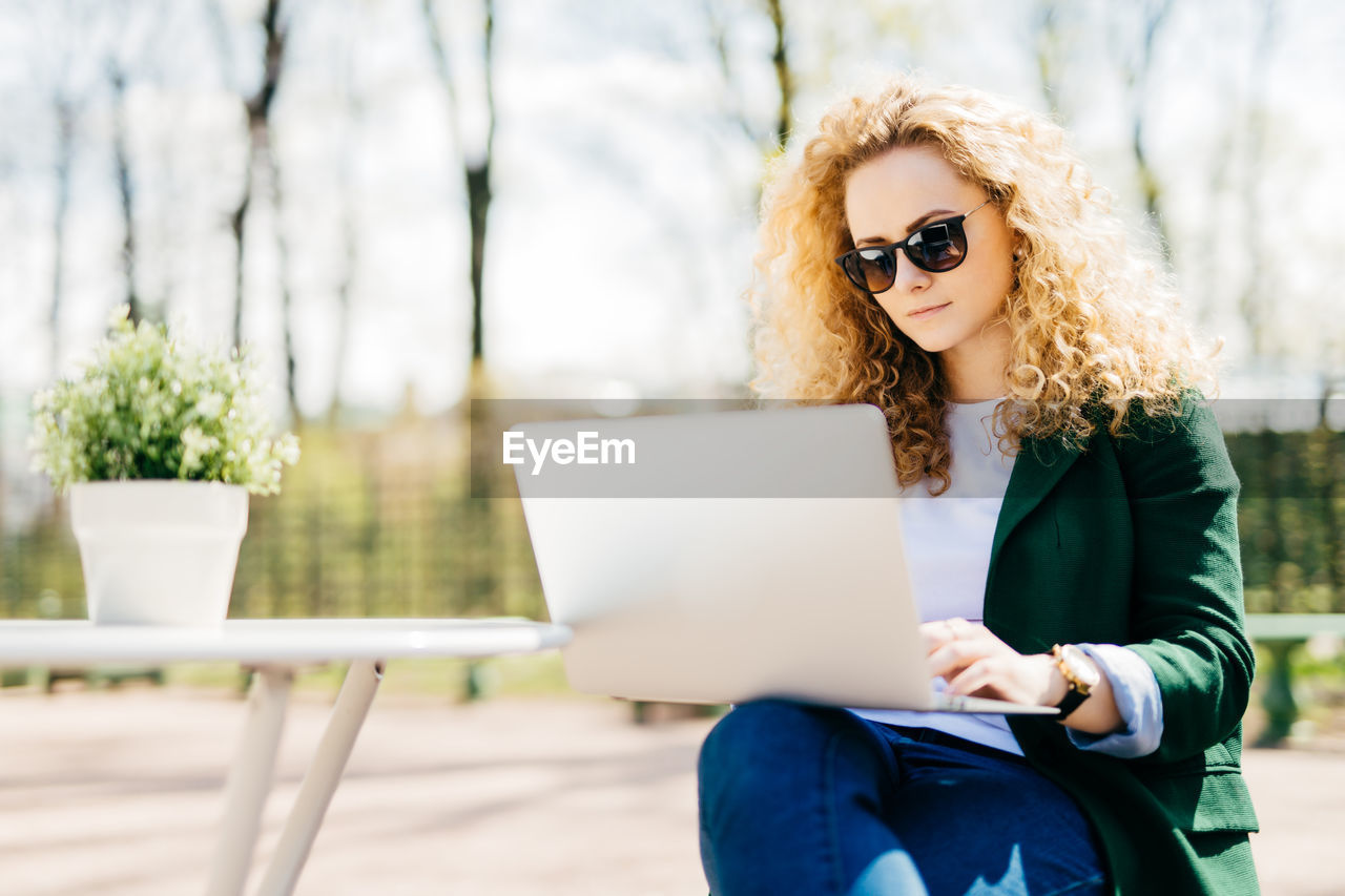 YOUNG WOMAN USING MOBILE PHONE WHILE SITTING ON TABLE OUTDOORS