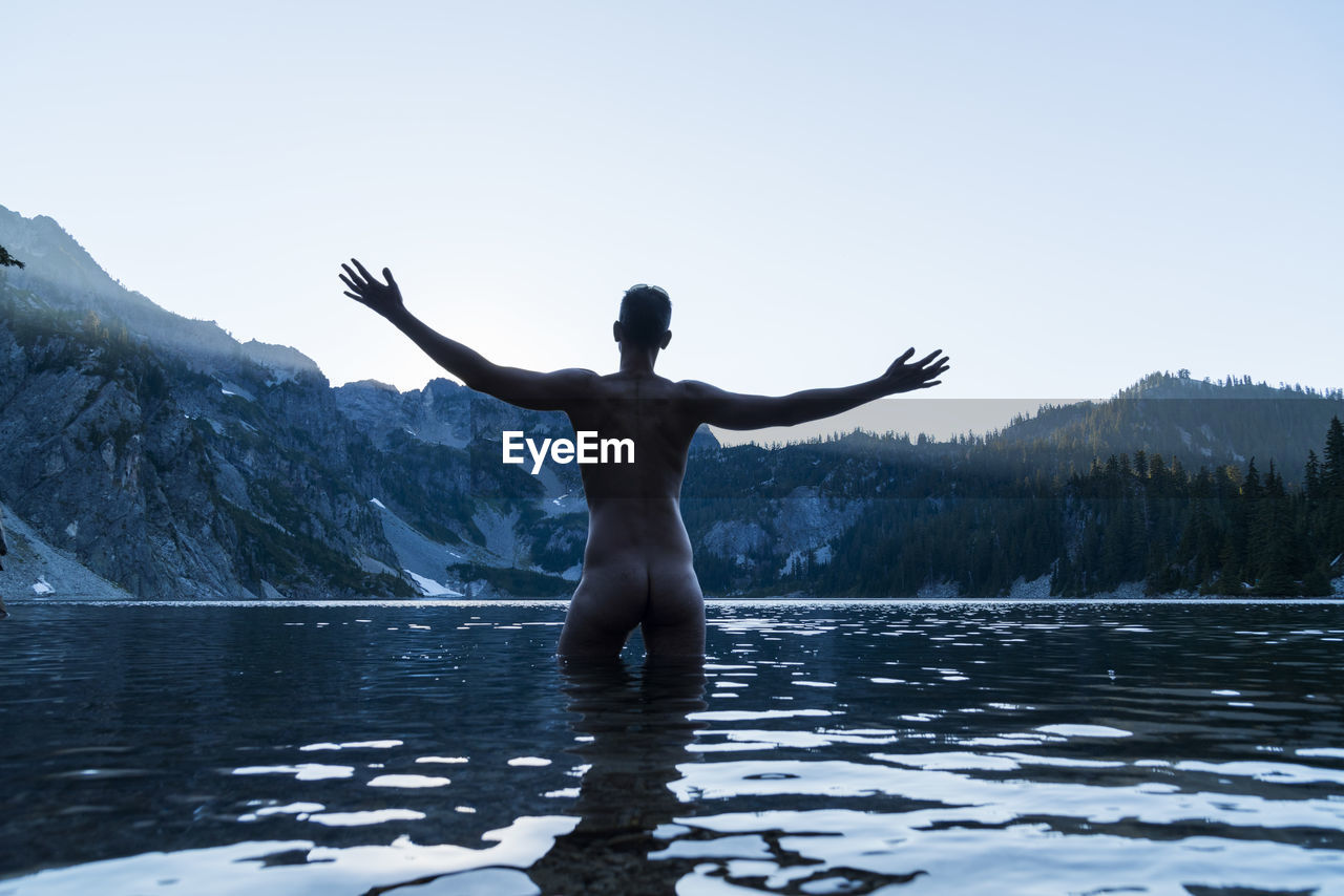 Rear view of naked man standing with arms outstretched in lake against clear sky
