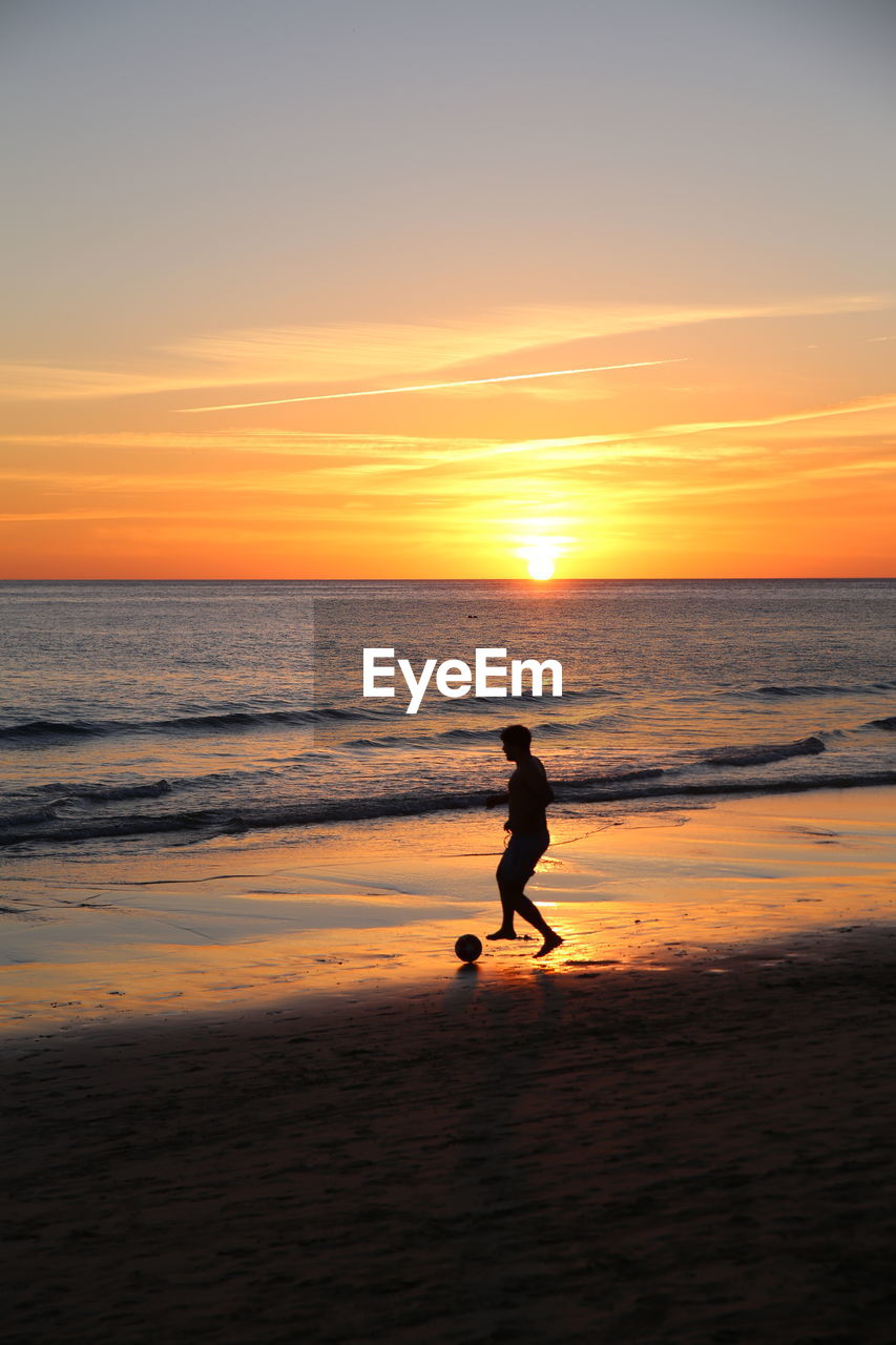 Full-length captured silhouette of a man playing football on the beach in front of sunset