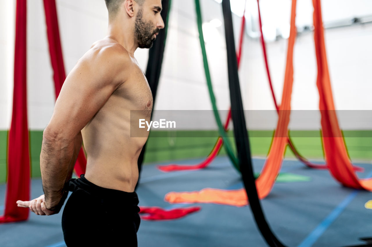 Side view of man in leotard bending and stretching arms while standing near colorful aerial silks during rehearsal