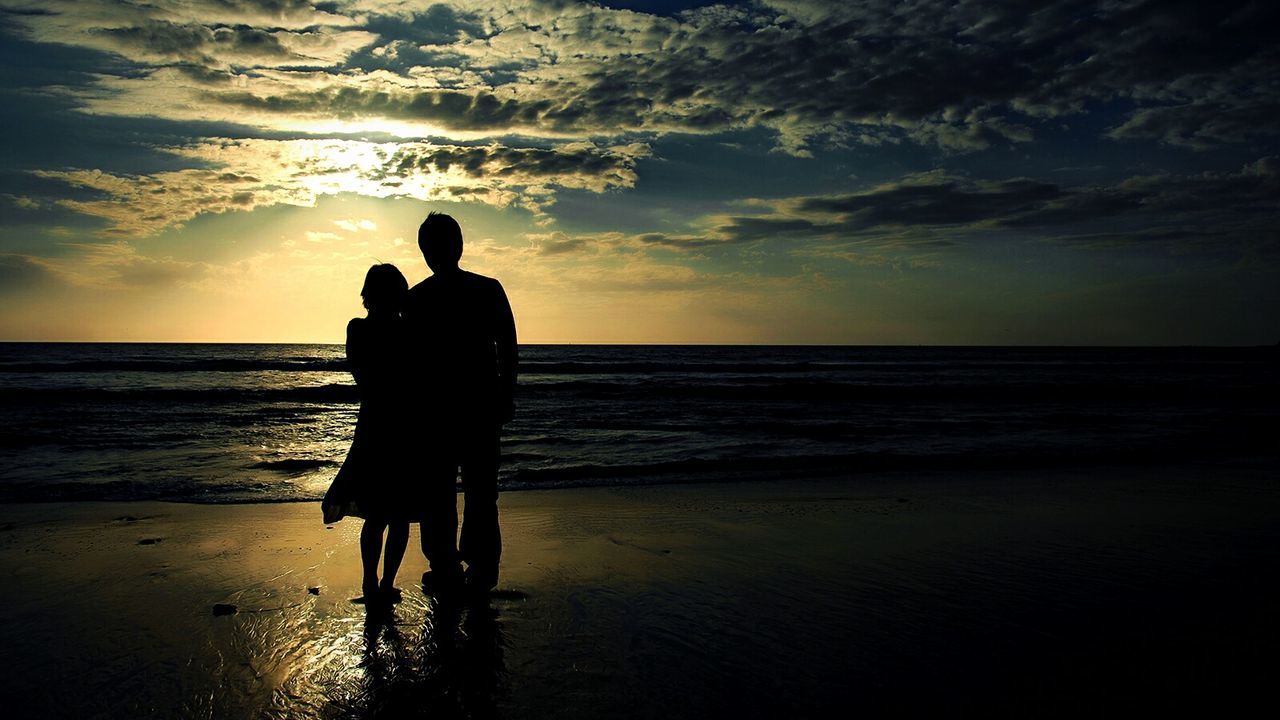 Rear view of silhouette man and woman standing on shore during sunset