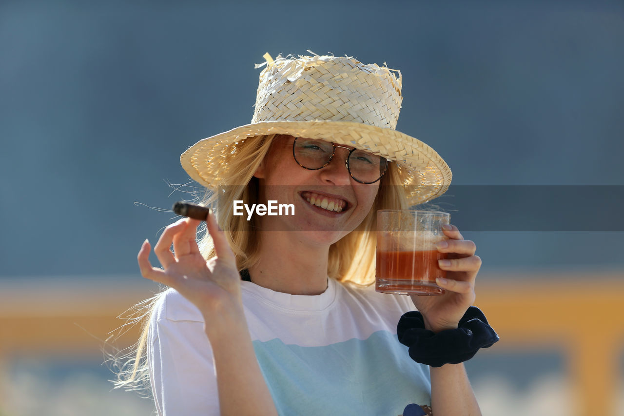 PORTRAIT OF A SMILING YOUNG WOMAN HOLDING HAT