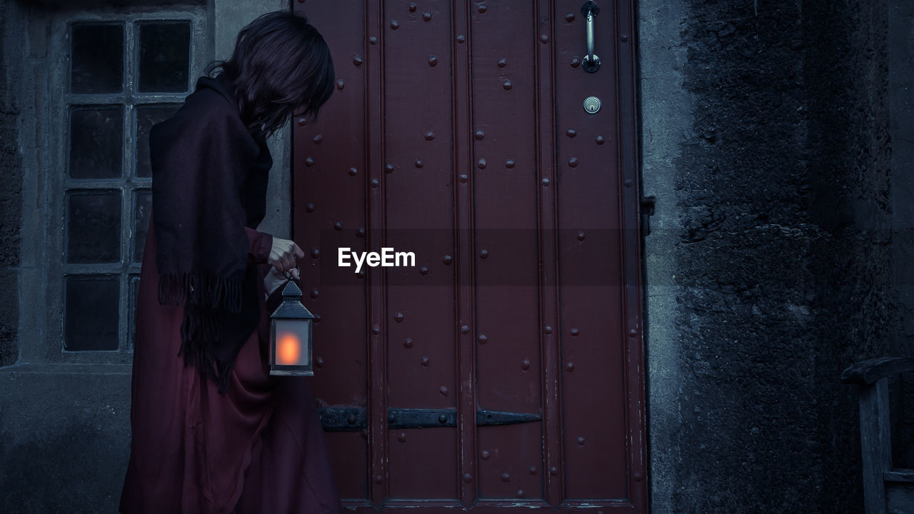 Side view of woman standing against closed door holding a light lantern