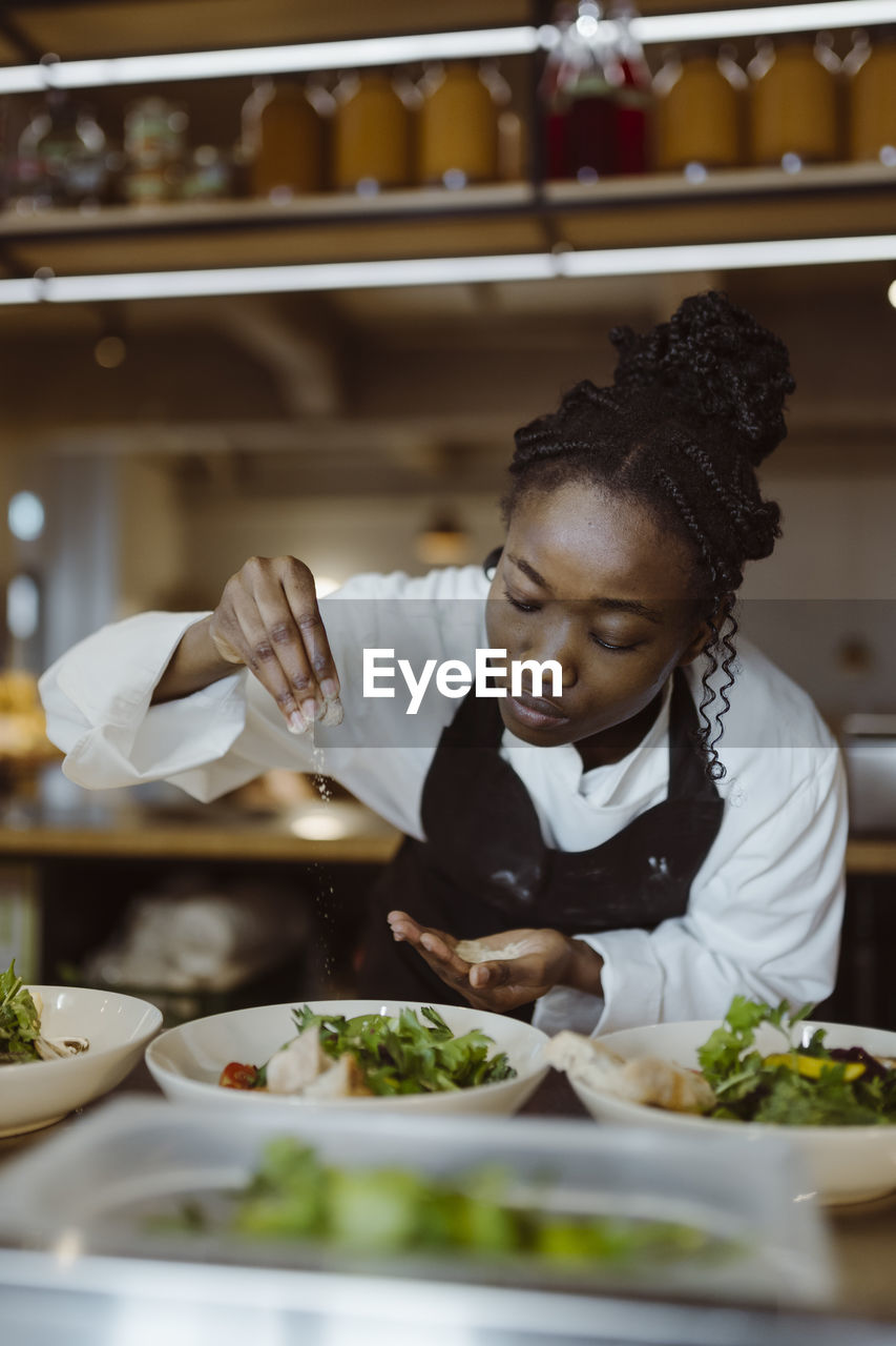 Focused young female chef sprinkling salt on salad while bending in commercial kitchen at restaurant