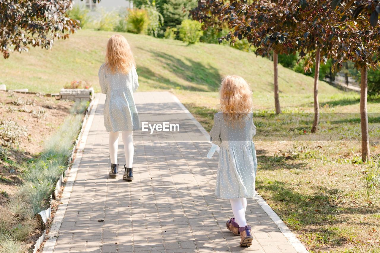 2 little blonde girls with curly hair are walking in a summer park.