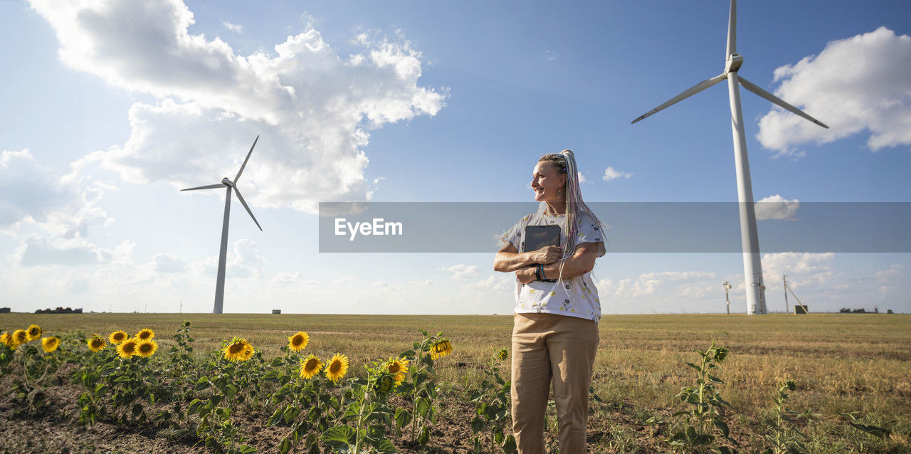 Young smiling woman with tablet in field with sunflowers, wind turbines for green energy produce,eco