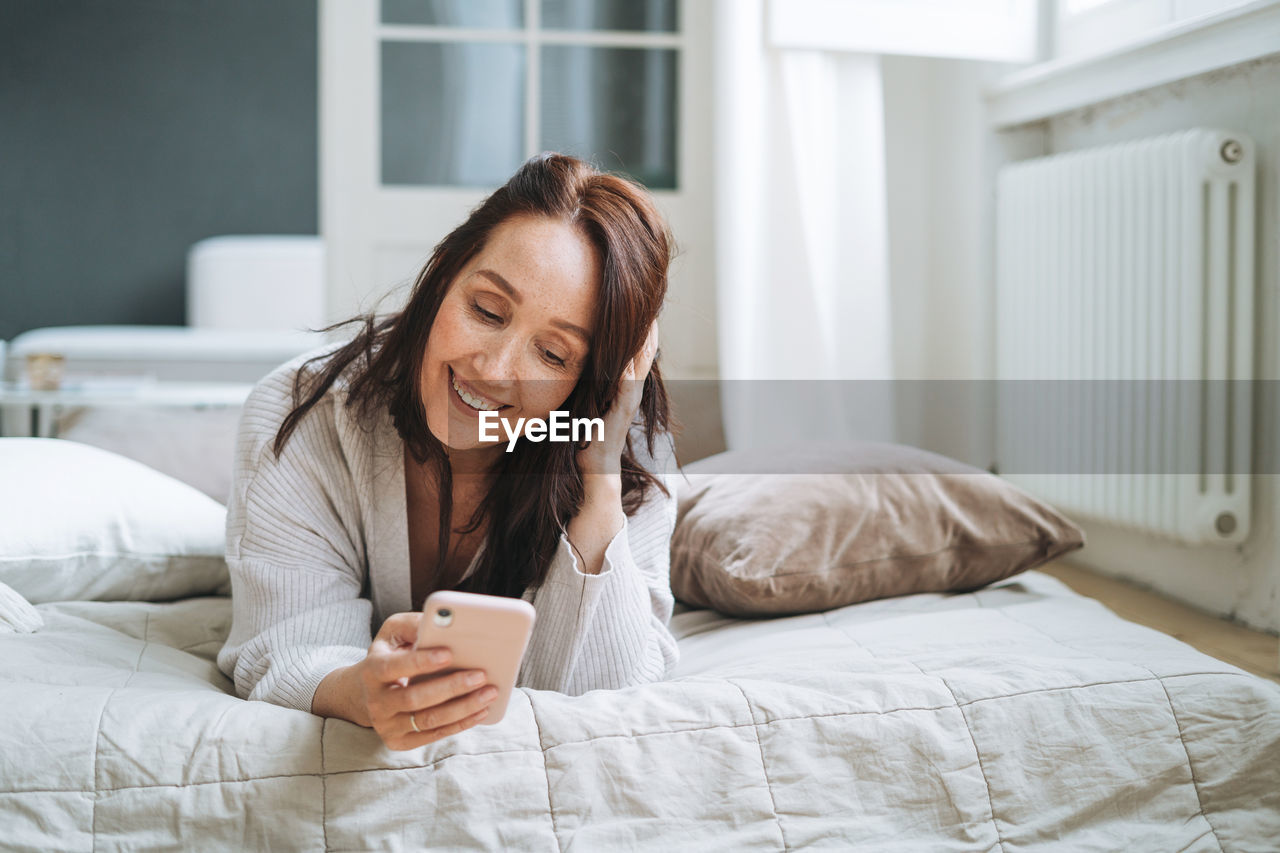 Young woman with brunette long hair in cozy knitted cardigan using mobile phone in bed at home