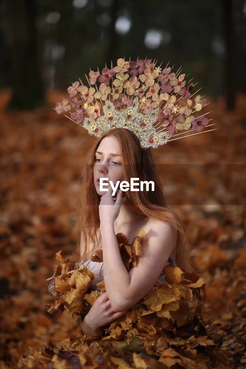 Thoughtful young woman covered with leaves sitting at forest during autumn