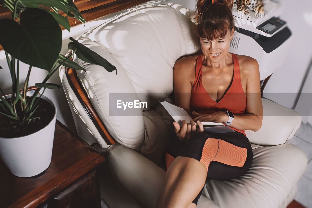 High angle view of smiling woman reading book while relaxing on chair at home