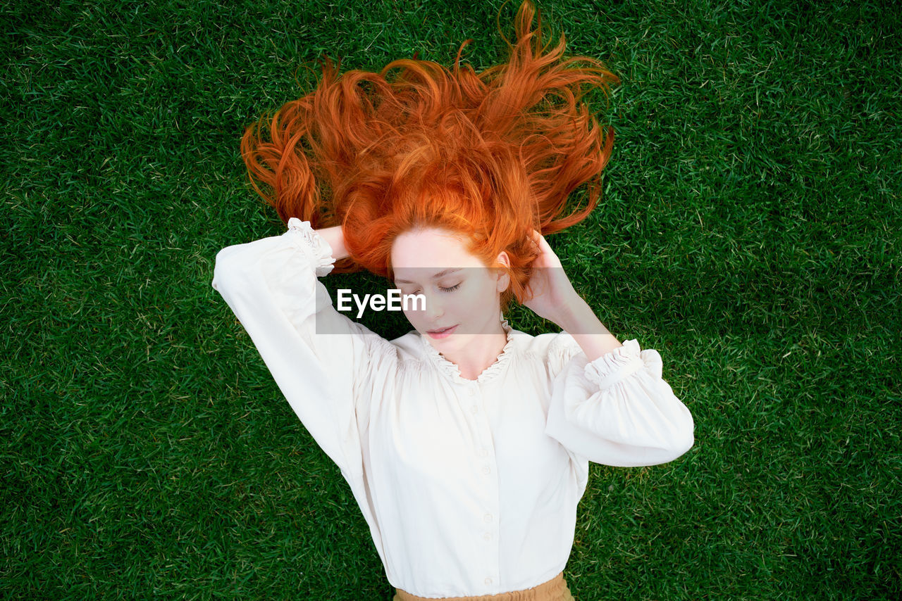 Redhead woman is resting on bright green lawn, her eyes closed and dreaming