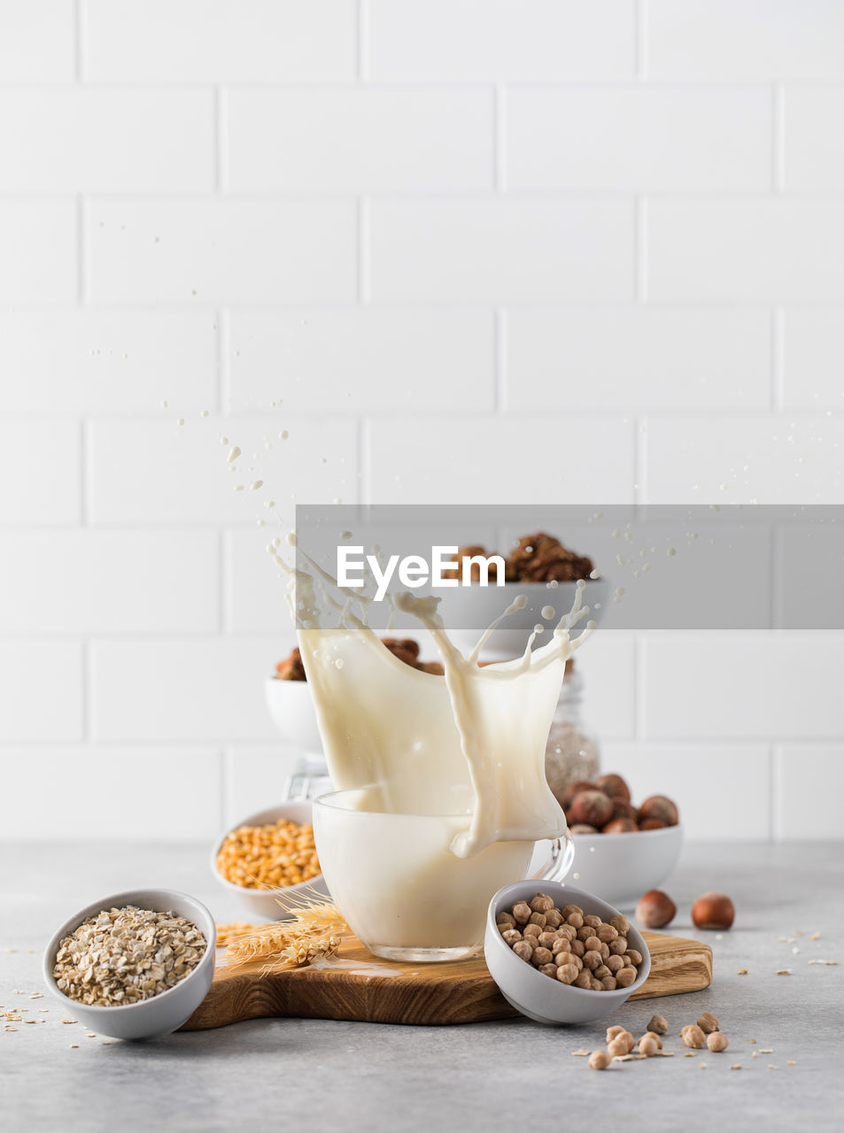 A splash of vegetable milk in a mug against the background of bowls with nuts, oatmeal and legumes.