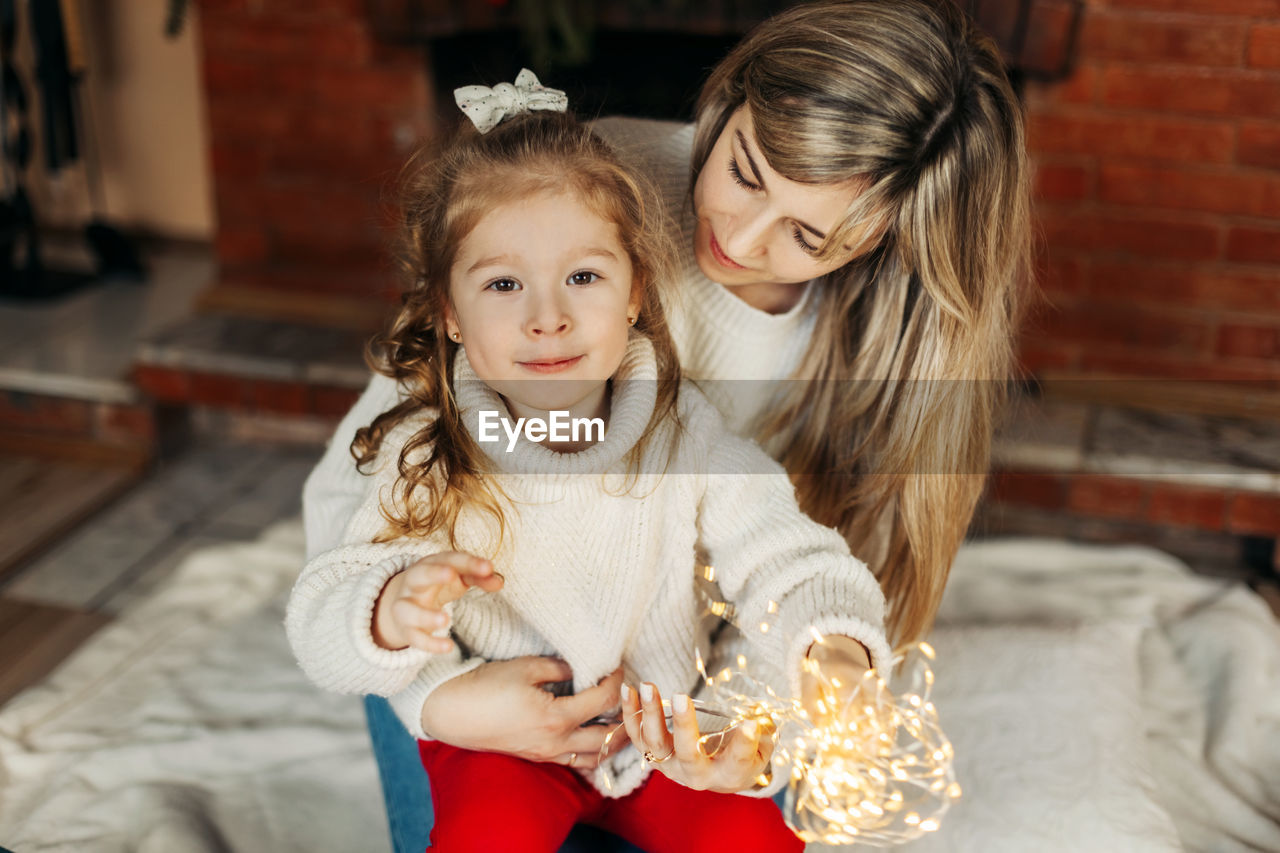 Mom and daughter are playing with christmas garlands on the floor by the fireplace
