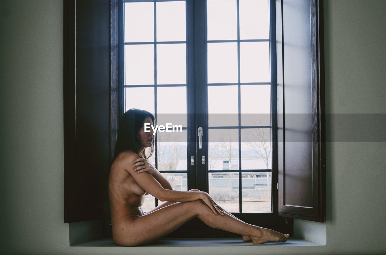 Side view of sensuous naked female model sitting on window sill