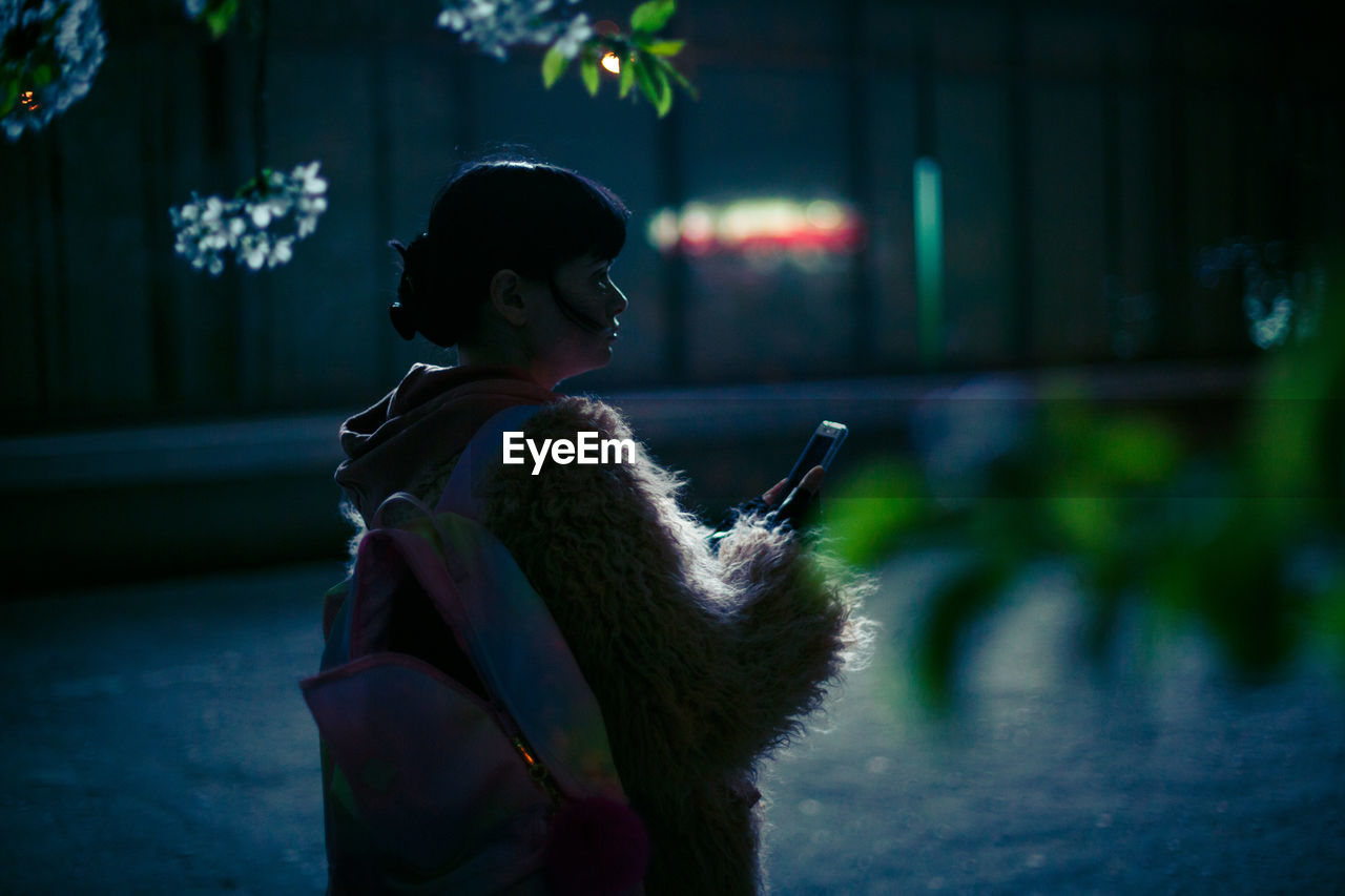 Woman holding phone while standing outdoors at night