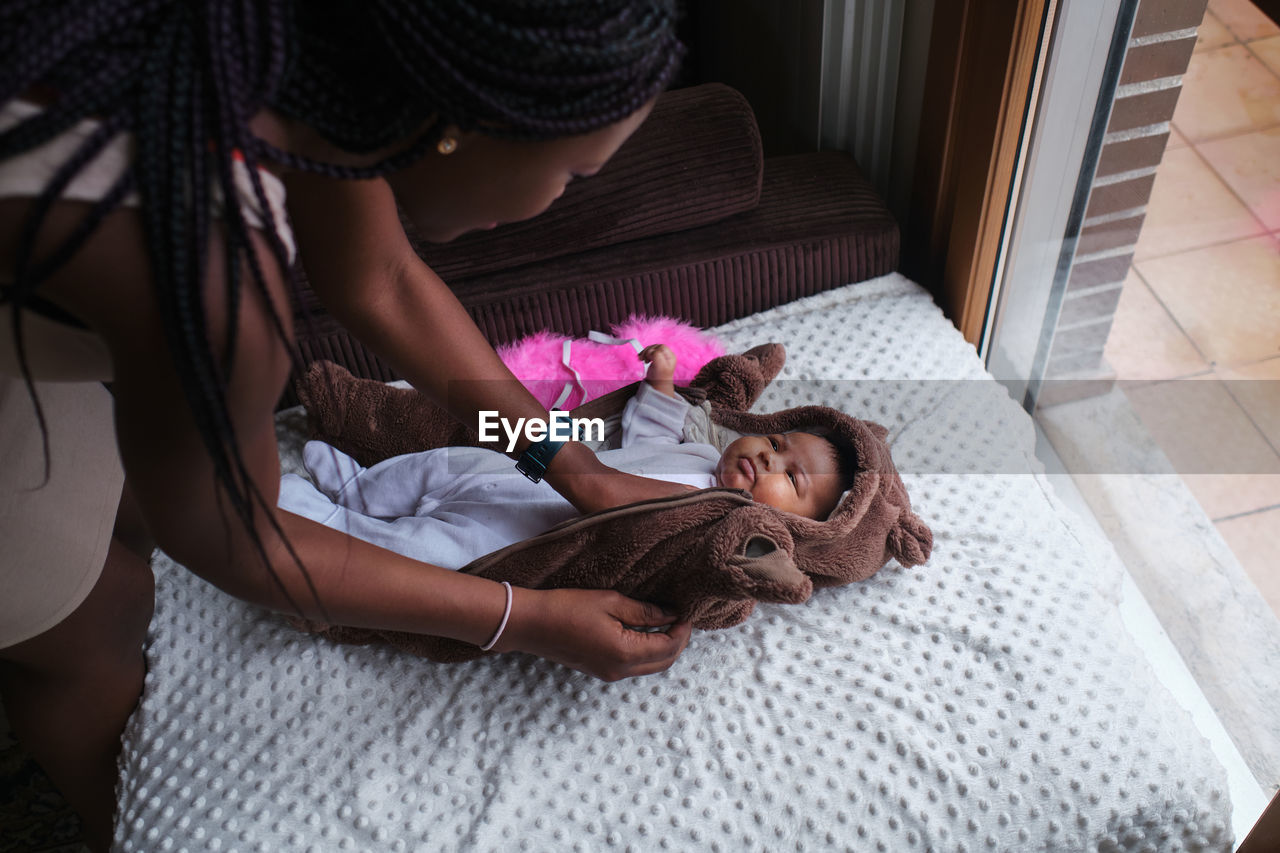 Caring african american mother with long braided hair dressing bodysuit on newborn baby lying on blanket on sofa