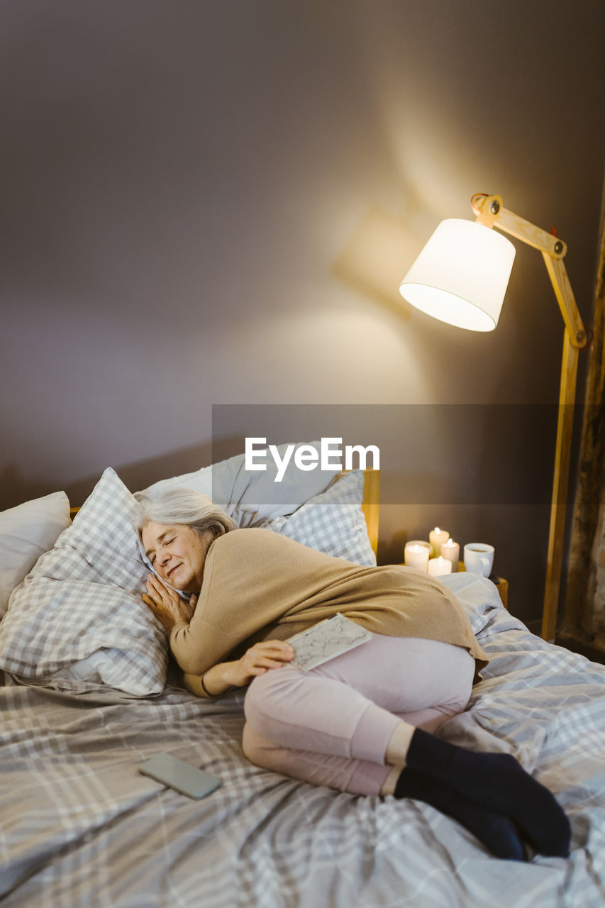 Senior woman sleeping on bed near illuminated lamp at home