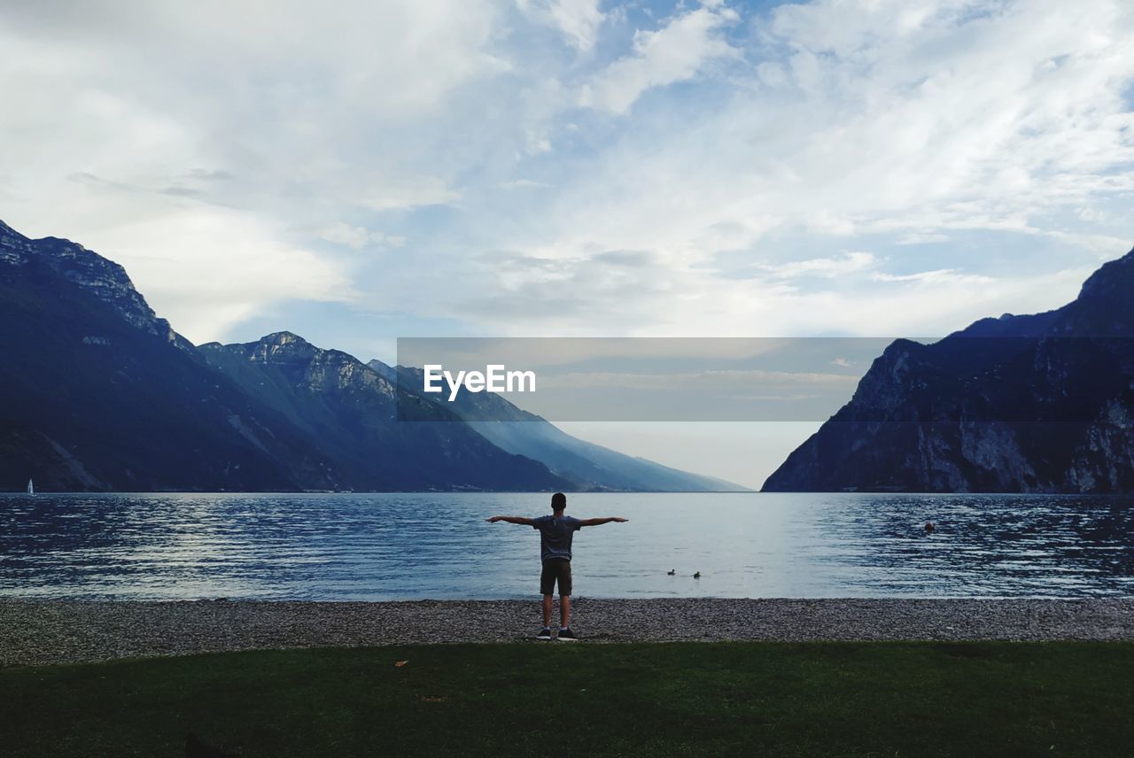 Rear view of boy with arms outstretched at lake against sky