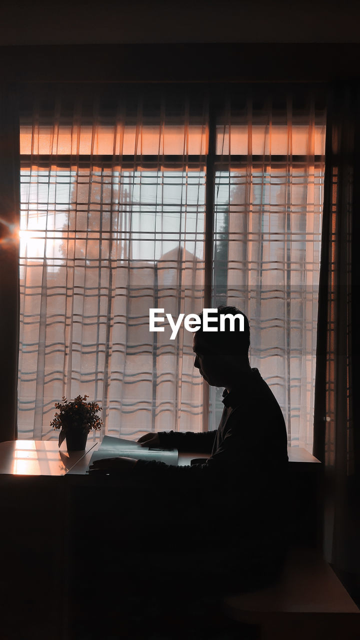 Side view of silhouette man reading book while sitting by window at home