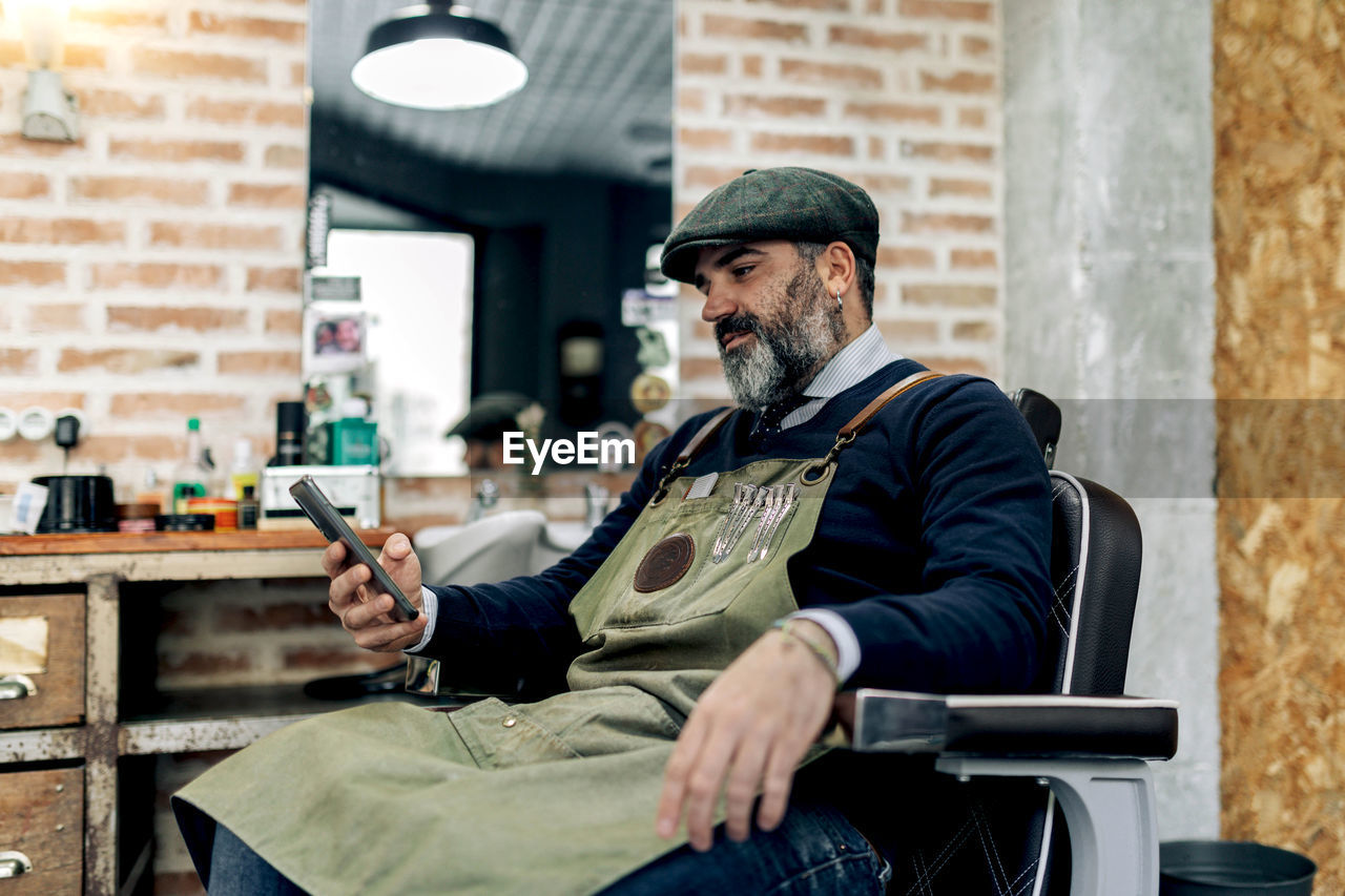 Side view of middle aged male barber sitting on chair in barbershop and browsing mobile phone while having break during work
