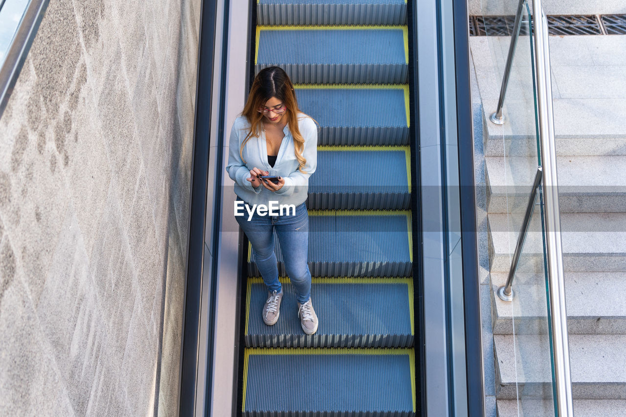 Cheerful lady with smartphone walking down on the escalator while smiling, photo from above