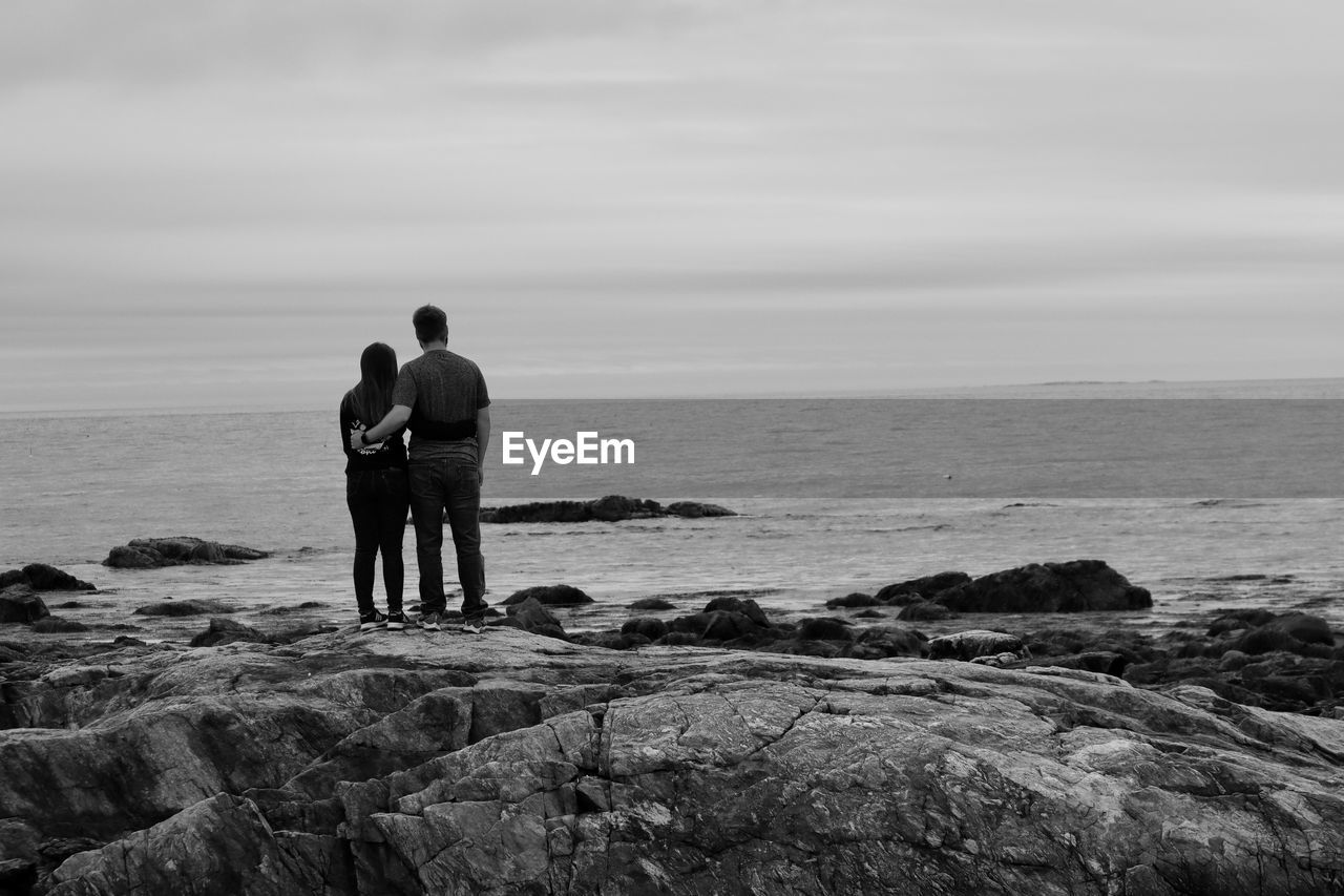 Rear view of engaged couple  standing on rock by sea against sky