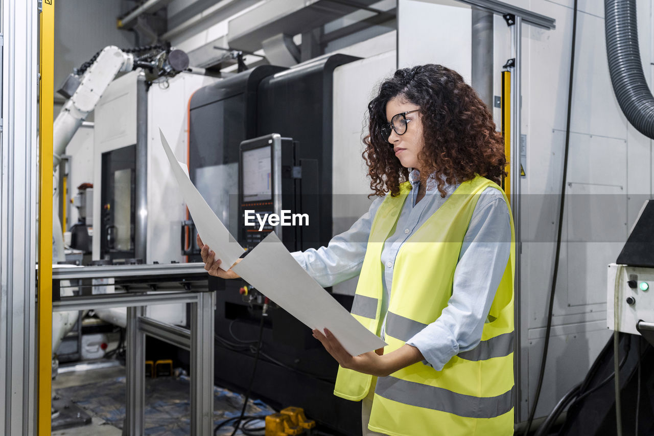 Young engineer examining blueprints standing by machine in industry