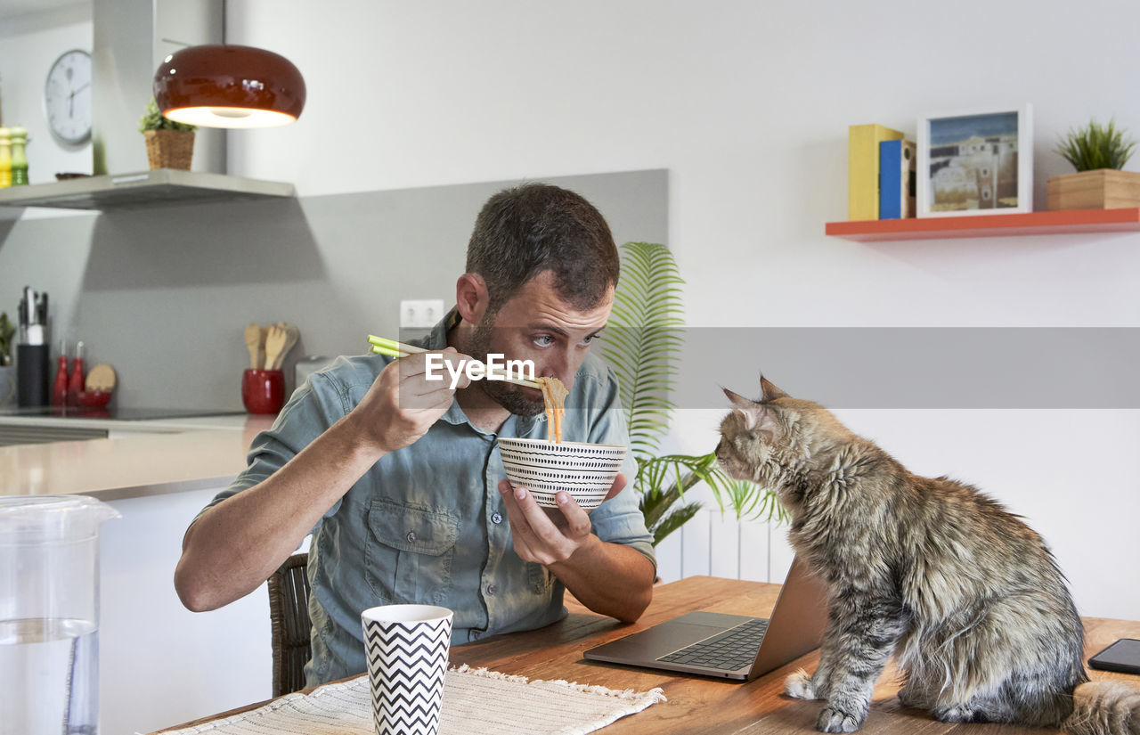 Male freelancer showing noodles to cat sitting on dining table at home