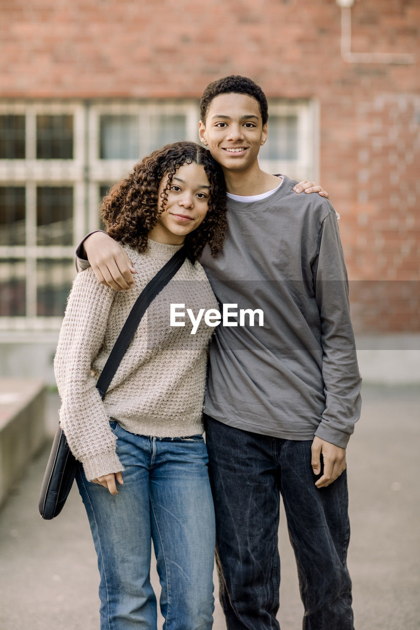 Portrait of smiling male and female teenage student standing with arms around in high school campus