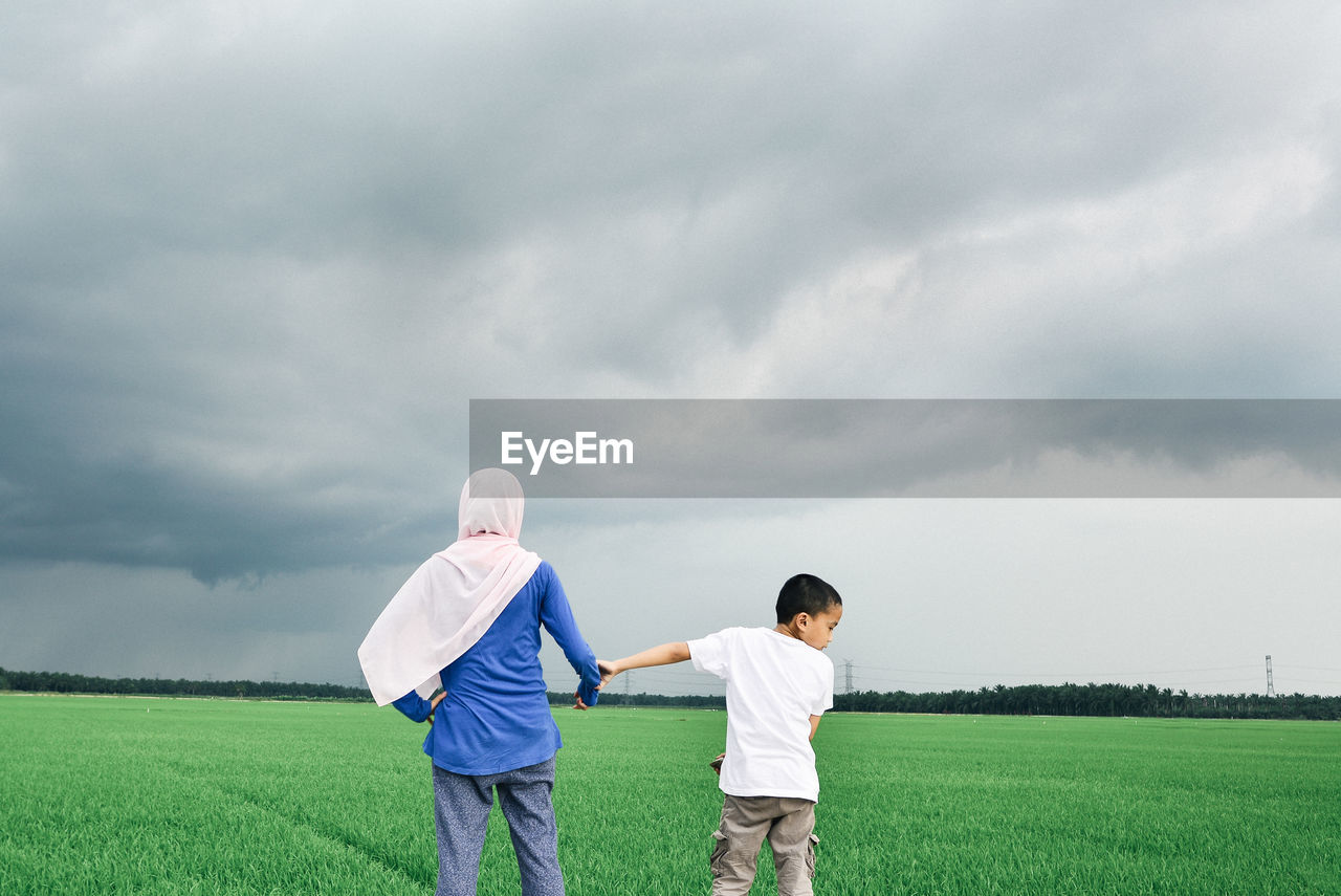 Woman wearing hijab standing with son on grassy field against storm clouds