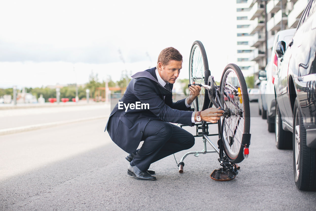 Businessman fixing bicycle chain on city street against sky