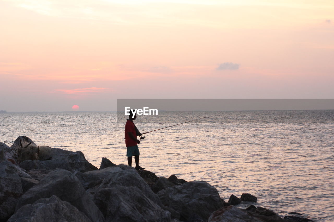 MAN STANDING ON ROCK AT BEACH DURING SUNSET