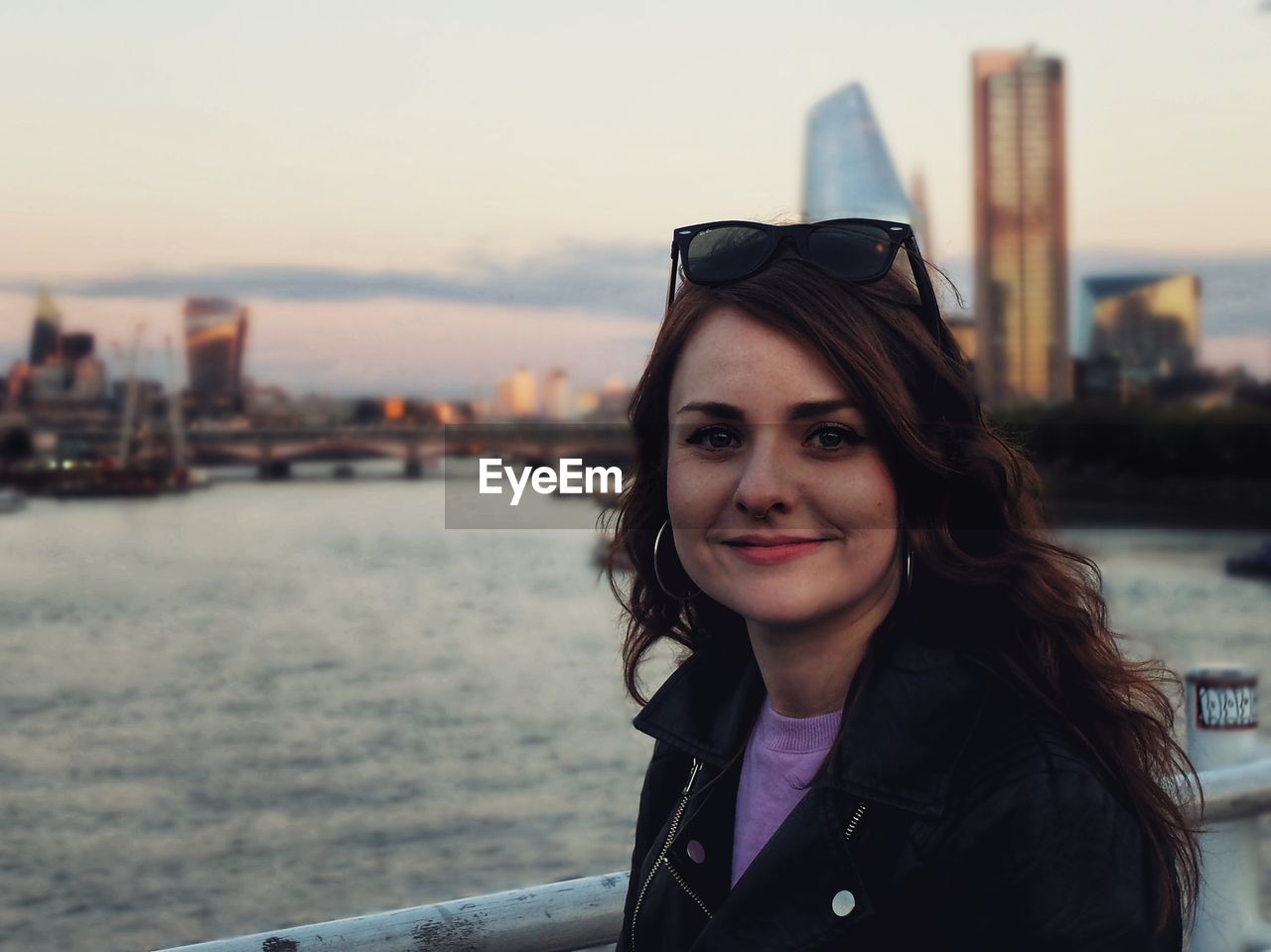 PORTRAIT OF SMILING YOUNG WOMAN IN CITY AGAINST SKY