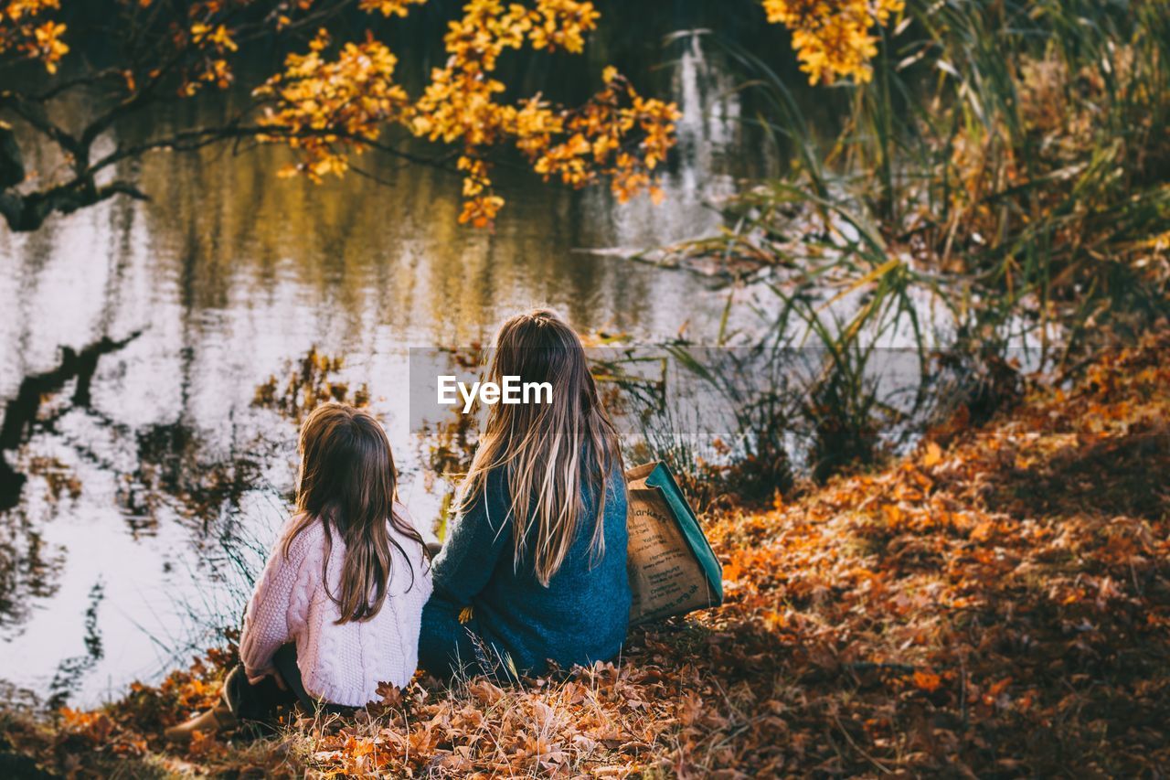 Rear view of women and daughter sitting by a pond during autumn