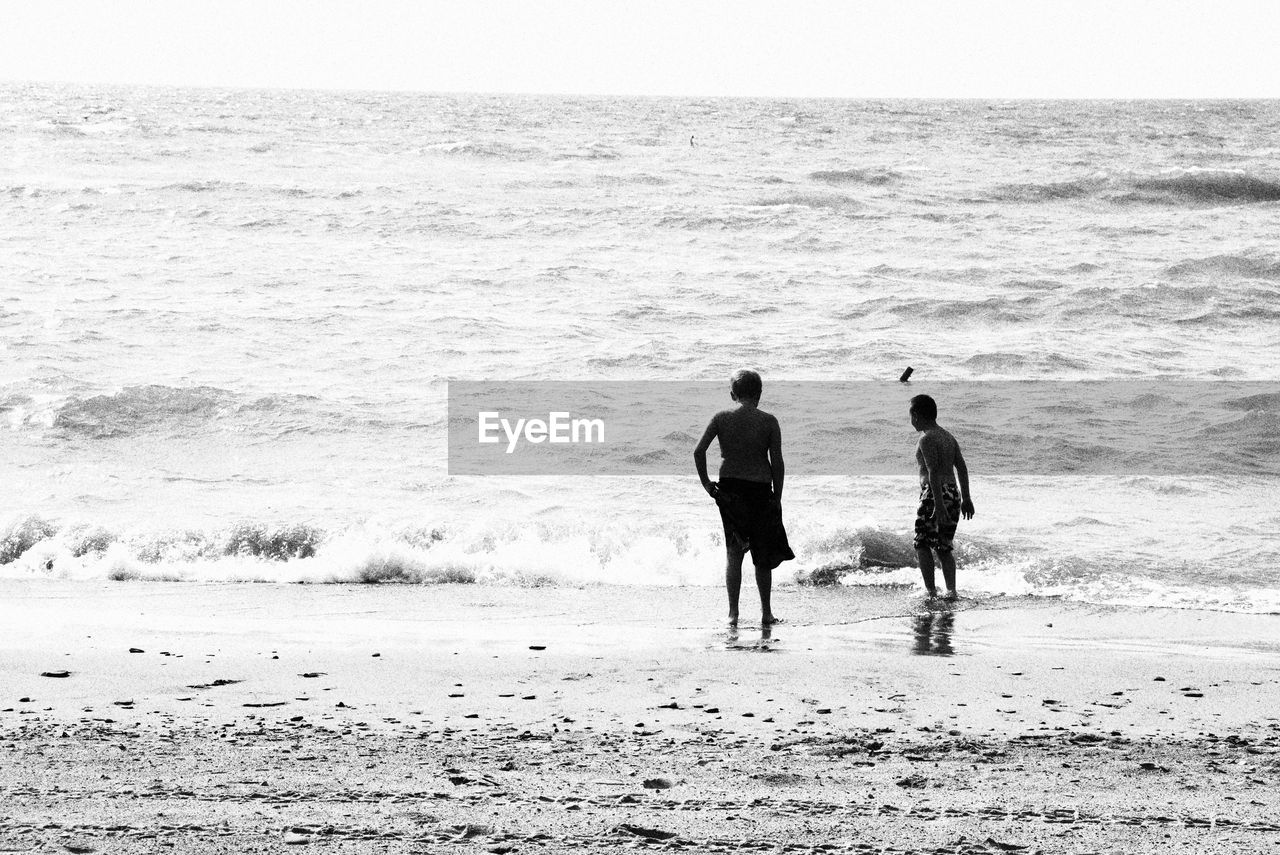 REAR VIEW OF MEN ON SHORE AT BEACH AGAINST SKY