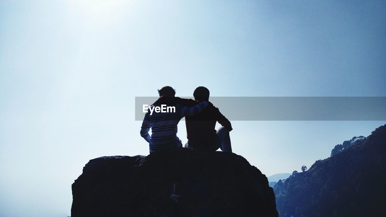 Rear view of men sitting on rock against clear sky during sunny day