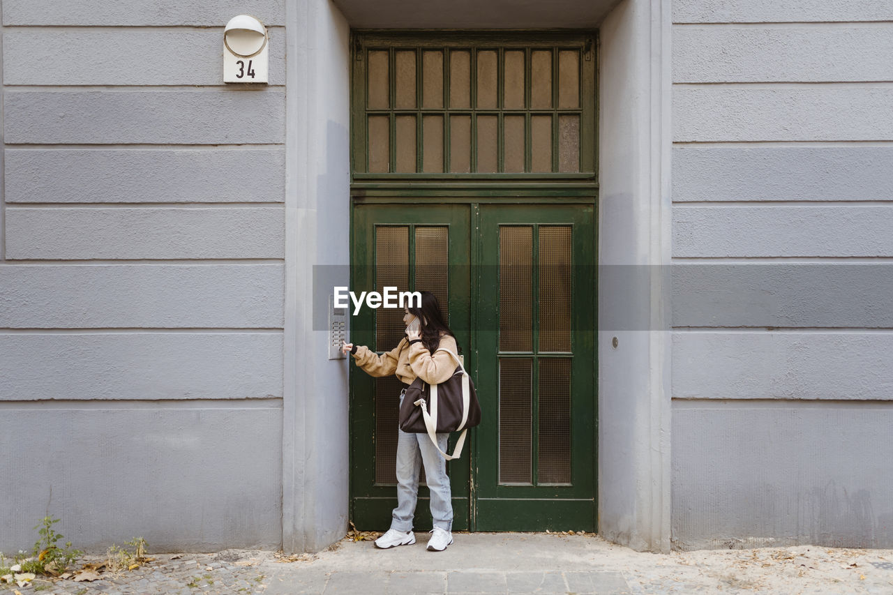 Woman talking on smart phone while doing intercom standing at apartment entrance