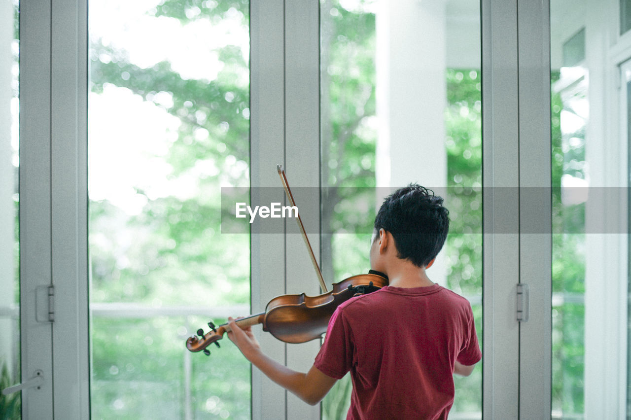 Rear view of boy playing violin by window