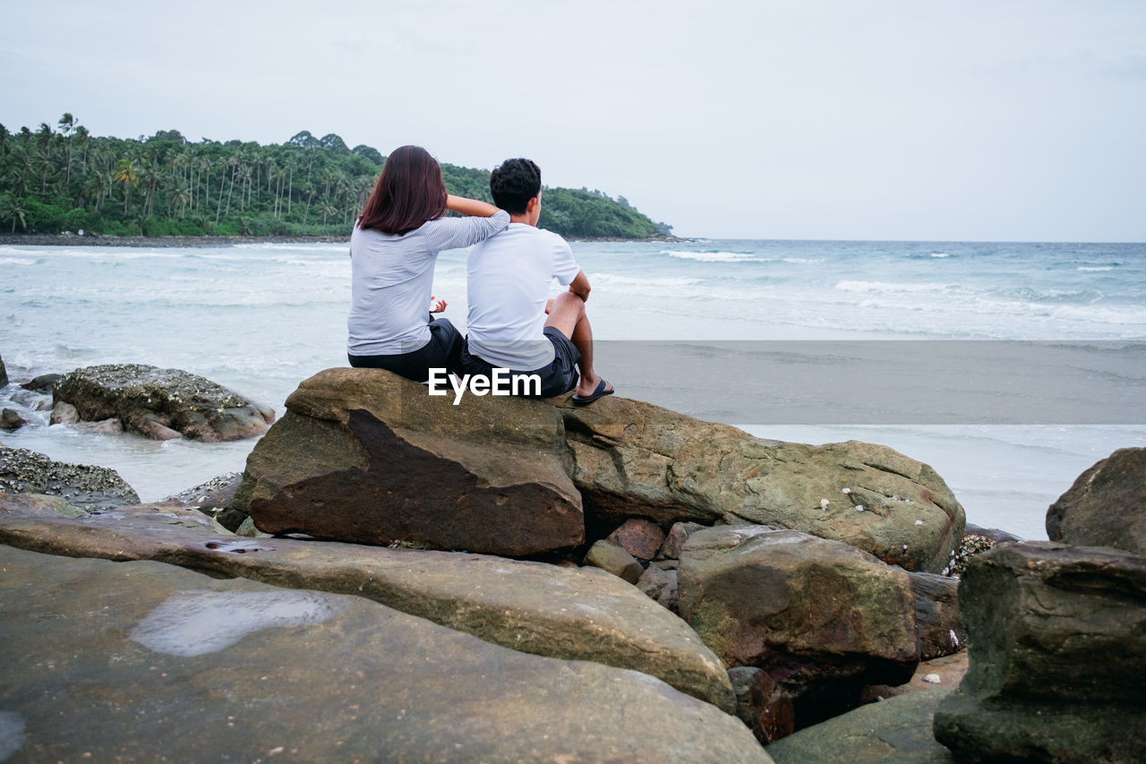 Rear view of couple sitting on rock against sea
