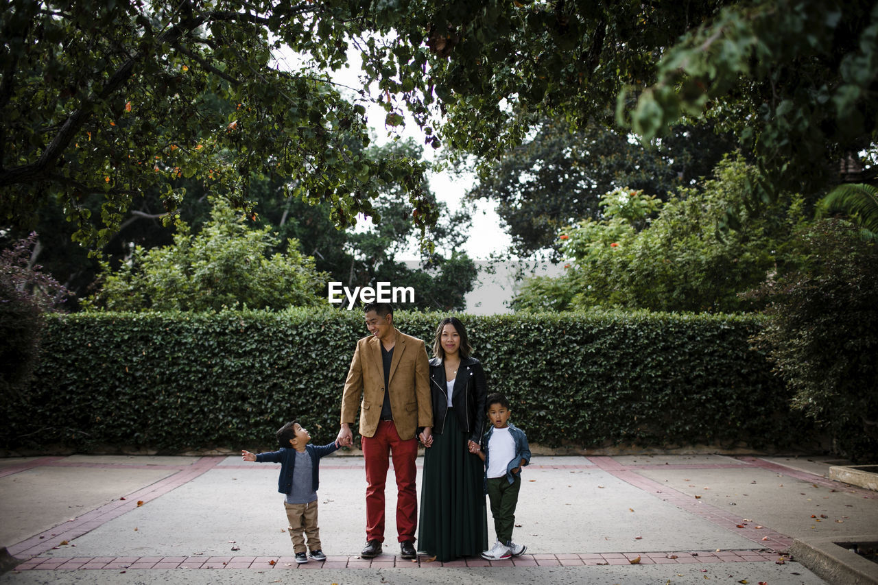 Smiling parents holding hands of cute sons while standing on footpath against plants at balboa park