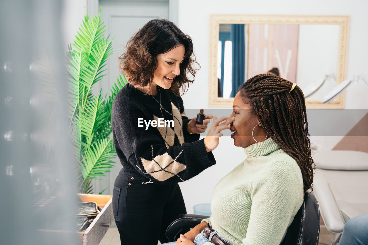 Focused female visagiste using fingers to smear foundation on cheek of black woman with braided hair during makeup session in studio