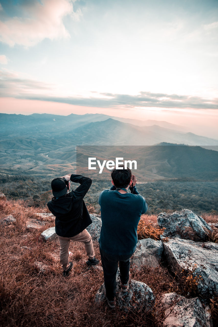 Rear view of friends photographing through camera while standing on mountain against sky during sunset