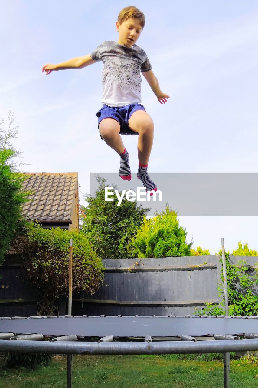 Full length of boy jumping on trampoline in back yard against sky