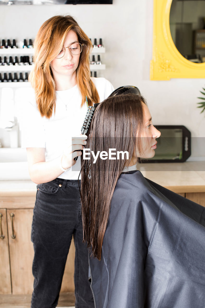 The hairdresser dries the client's hair with a hair dryer and styles her hair with a comb.