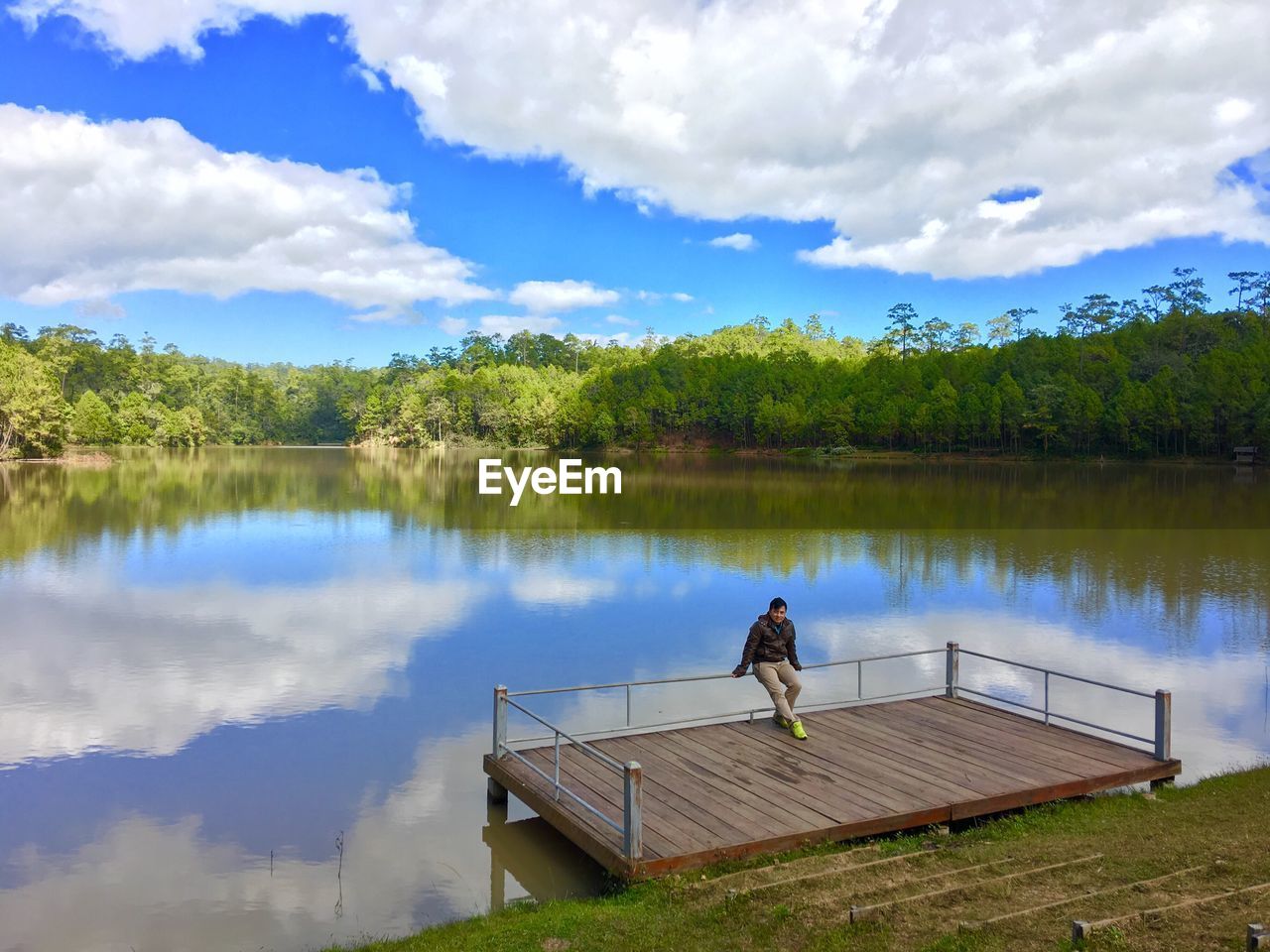 Man sitting on railing of floorboard over lake against sky