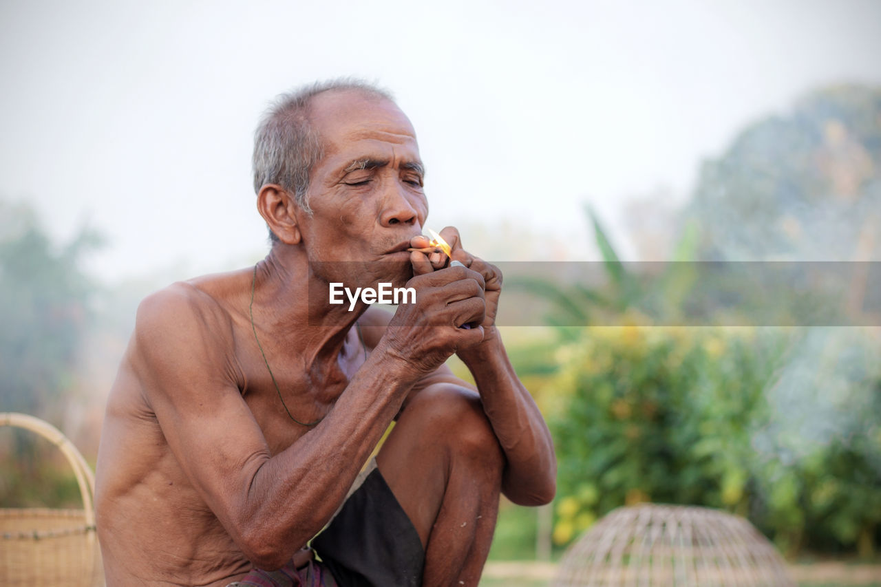 Shirtless senior man lighting bidi while sitting at farm