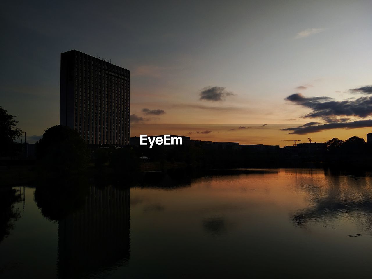 Silhouette buildings by lake against sky during sunset
