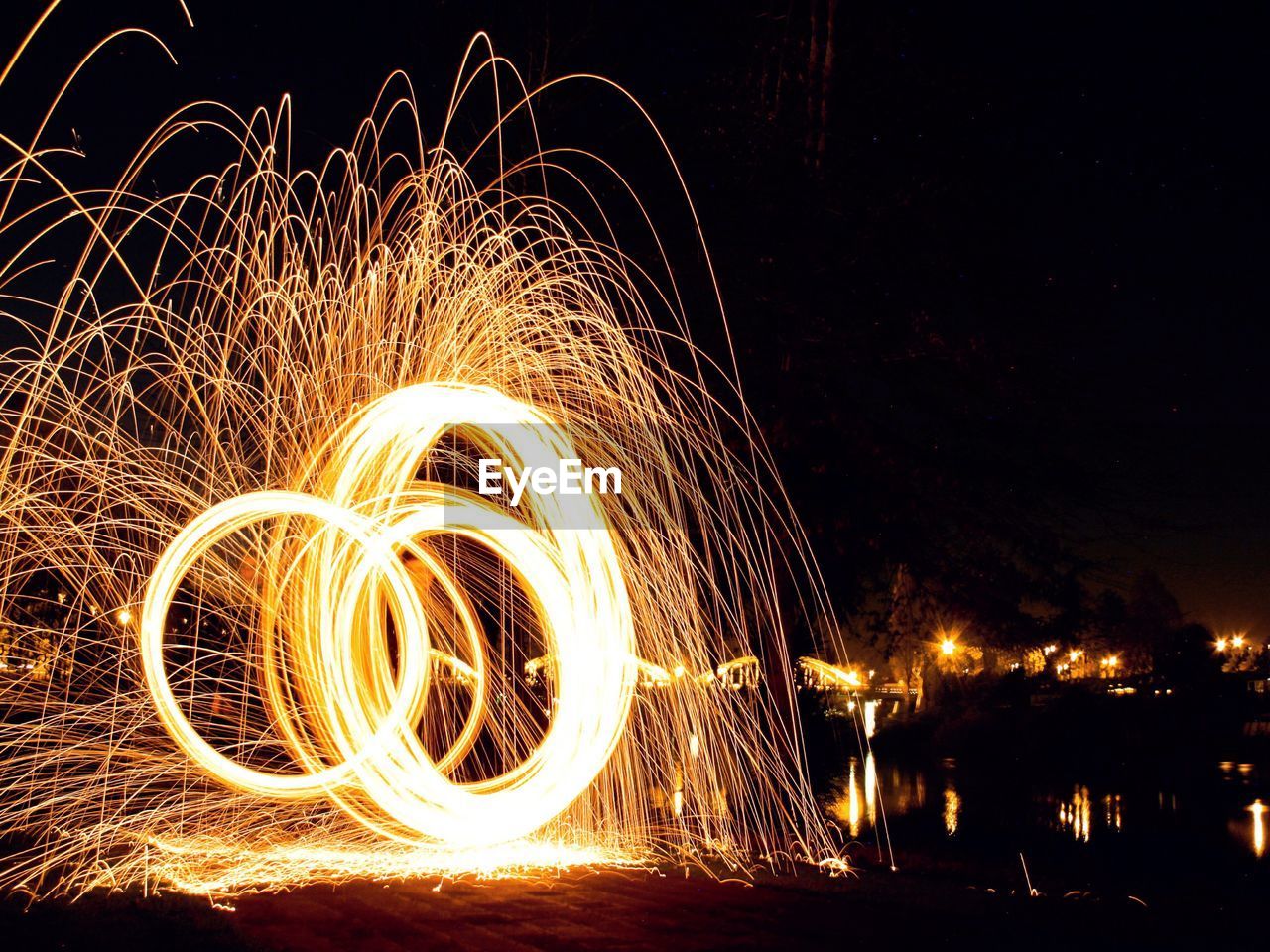 Illuminated wire wool in motion at riverbank against clear sky at night