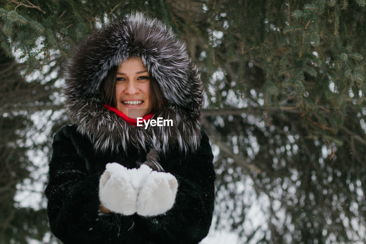 Asian girl in a black coat and fluffy fur hat smiles. she holds a handful of snow in white mittens. 