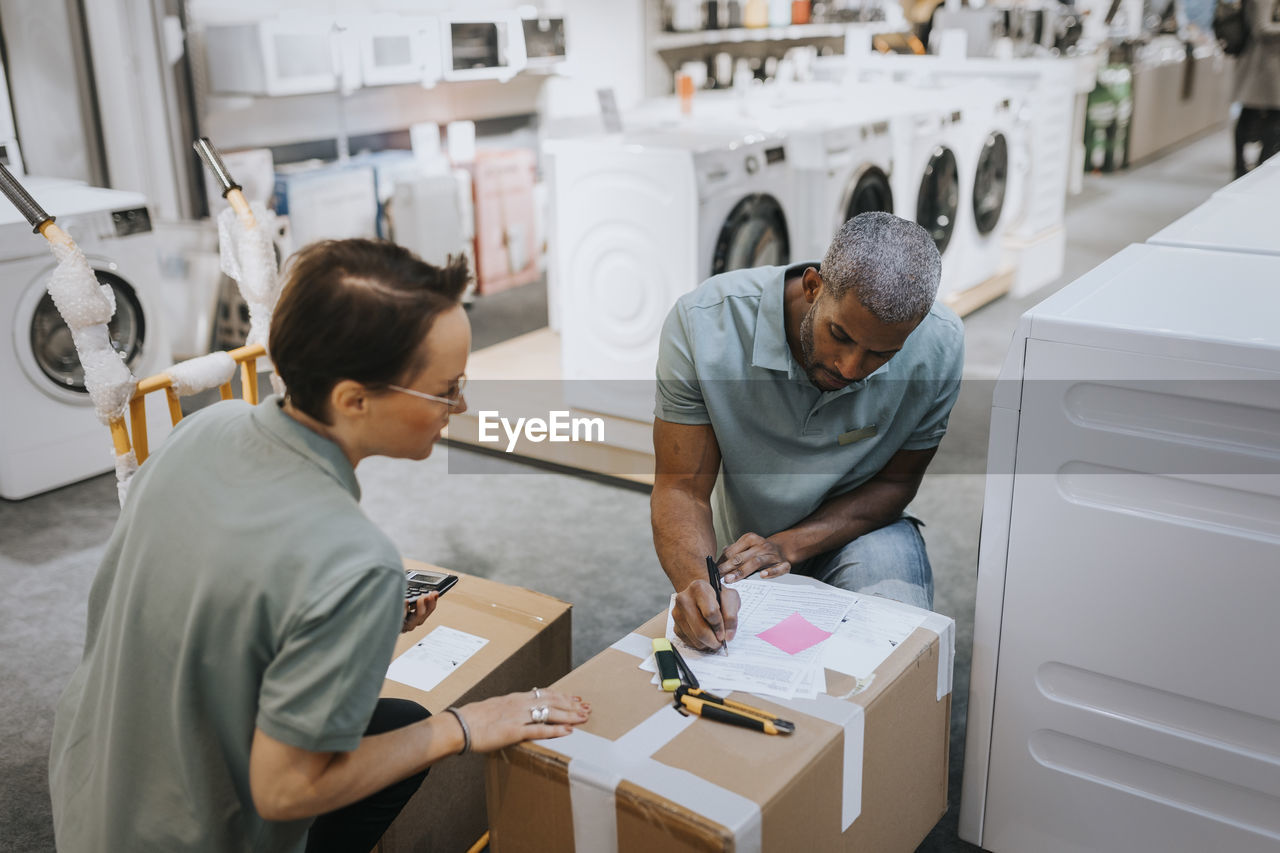 Saleswoman looking at salesman writing in document on box at appliances store